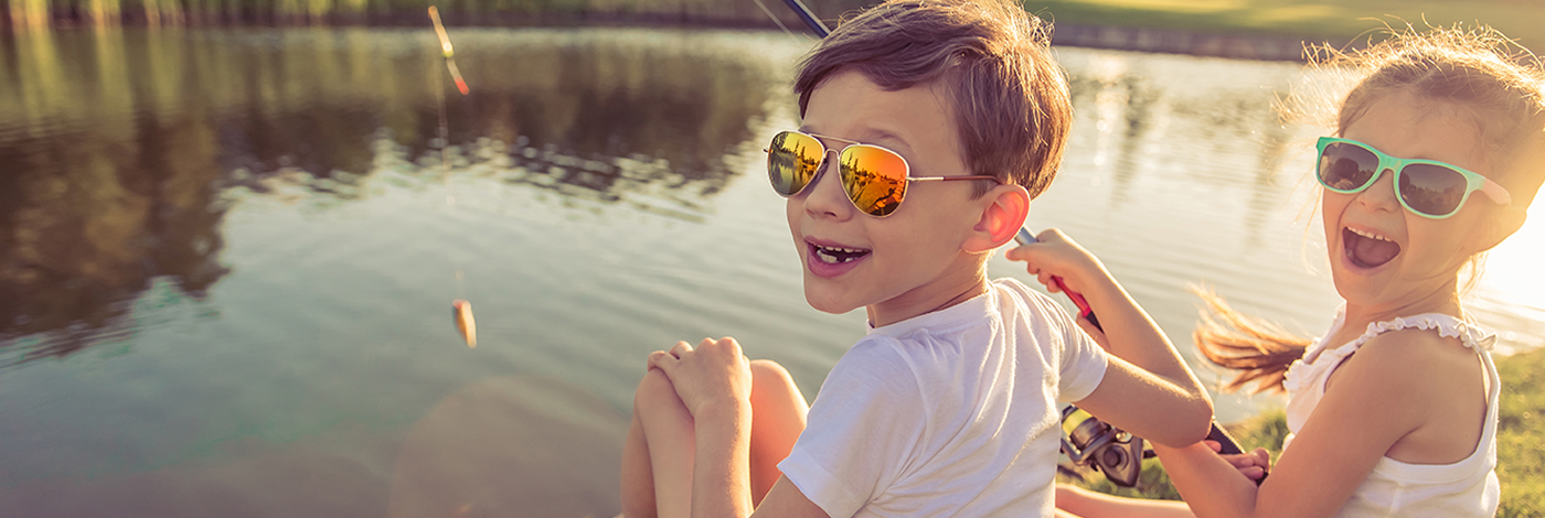 Funny stylish little boy and girl in sun glasses are looking at camera and smiling while catching fish in the pond using a fishing rod, sitting on the ground
